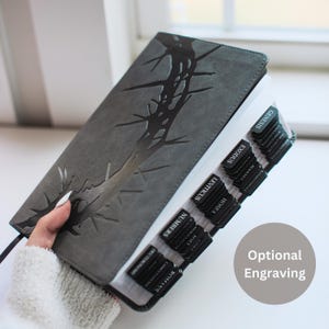 May include: Gray Bible with embossed design and black page tabs. The tabs are labeled with book names. The cover features a dark, textured pattern. The text "Optional Engraving" is visible.