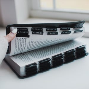 May include: A black Bible with labeled tabs for easy navigation. The open pages reveal text, with the tabs indicating sections like "2 Samuel," "Numbers," and "Leviticus." The book is held open, showcasing the interior pages.