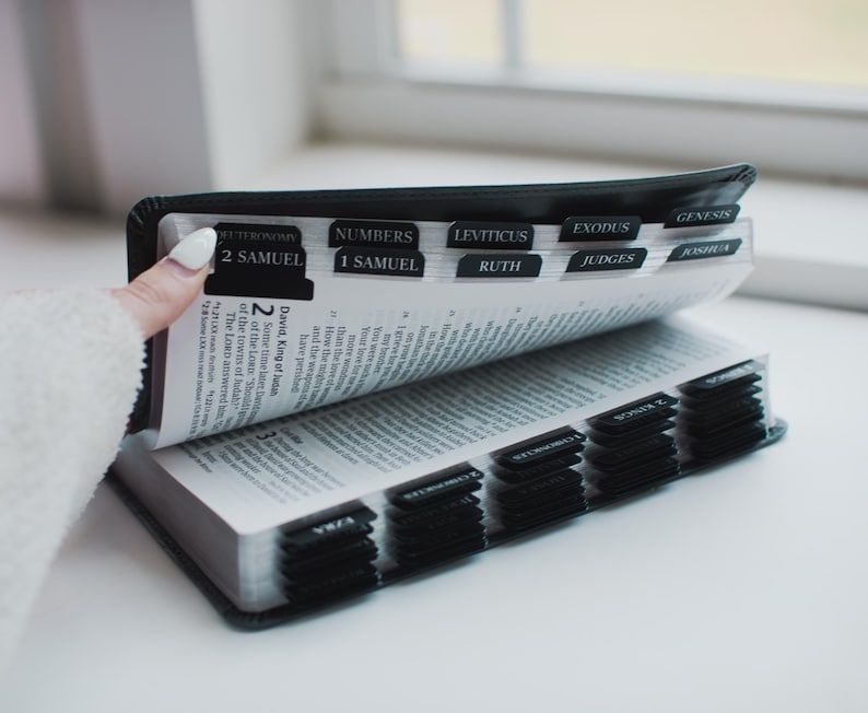 May include: A black Bible with labeled tabs for easy navigation. The open pages reveal text, with the tabs indicating sections like "2 Samuel," "Numbers," and "Leviticus." The book is held open, showcasing the interior pages.