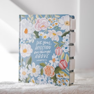 K&ouml;nnte beinhalten: Ein Notizbuch mit hellblauem Einband, verziert mit einem floralen Design in Wei&szlig;, Rosa und Orange. Der Einband zeigt den Text "Set your affection on things above". Das Notizbuch hat beschriftete Registerkarten.