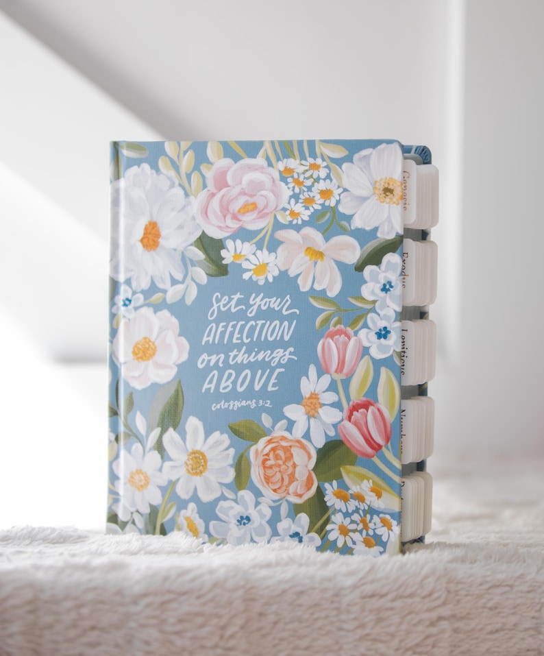 K&ouml;nnte beinhalten: Ein Notizbuch mit hellblauem Einband, verziert mit einem floralen Design in Wei&szlig;, Rosa und Orange. Der Einband zeigt den Text "Set your affection on things above". Das Notizbuch hat beschriftete Registerkarten.