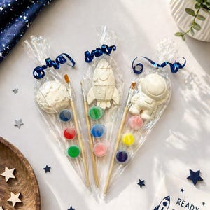 May include: Three space-themed paint kits in clear bags, each with a different shape: a planet, a rocket, and an astronaut. Each kit includes a paintbrush and a set of colourful paints. A card with the text "READY SET PAINT!" is also included.