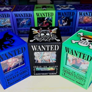 May include: A collection of rectangular storage boxes featuring One Piece Wanted posters. The front displays character portraits with the text WANTED, DEAD OR ALIVE, and character names. The boxes are in various colors including blue, black, and green.