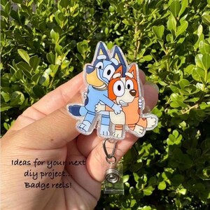 May include: A badge reel featuring a cartoon image of two hugging dogs, one blue and one orange. The badge reel has a clear plastic covering and a silver retractable clip. Text on the image reads "Ideas for your next diy project... Badge reels!"