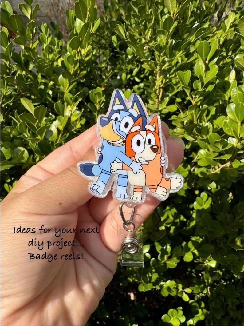 May include: A badge reel featuring a cartoon image of two hugging dogs, one blue and one orange. The badge reel has a clear plastic covering and a silver retractable clip. Text on the image reads "Ideas for your next diy project... Badge reels!"