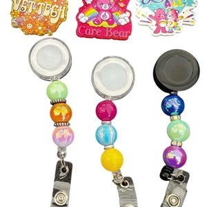 May include: Three badge reels with decorative beads and matching decorative tops. The reels have different designs, including a floral "VET TECH" badge, a Care Bear with the text "Don't Fucking", and a Care Bear with the text "Love All". The text "What will you create?" is at the top.