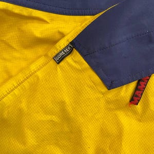 May include: A yellow and navy blue jacket featuring a grid-patterned fabric texture. A black Gore-Tex tag is stitched onto the seam, and a partial red Mammut brand label is visible on the lower right side of the navy blue panel section.