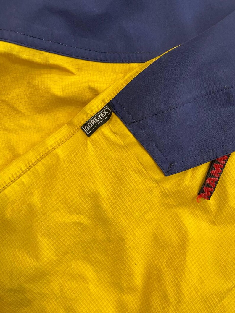 May include: A yellow and navy blue jacket featuring a grid-patterned fabric texture. A black Gore-Tex tag is stitched onto the seam, and a partial red Mammut brand label is visible on the lower right side of the navy blue panel section.