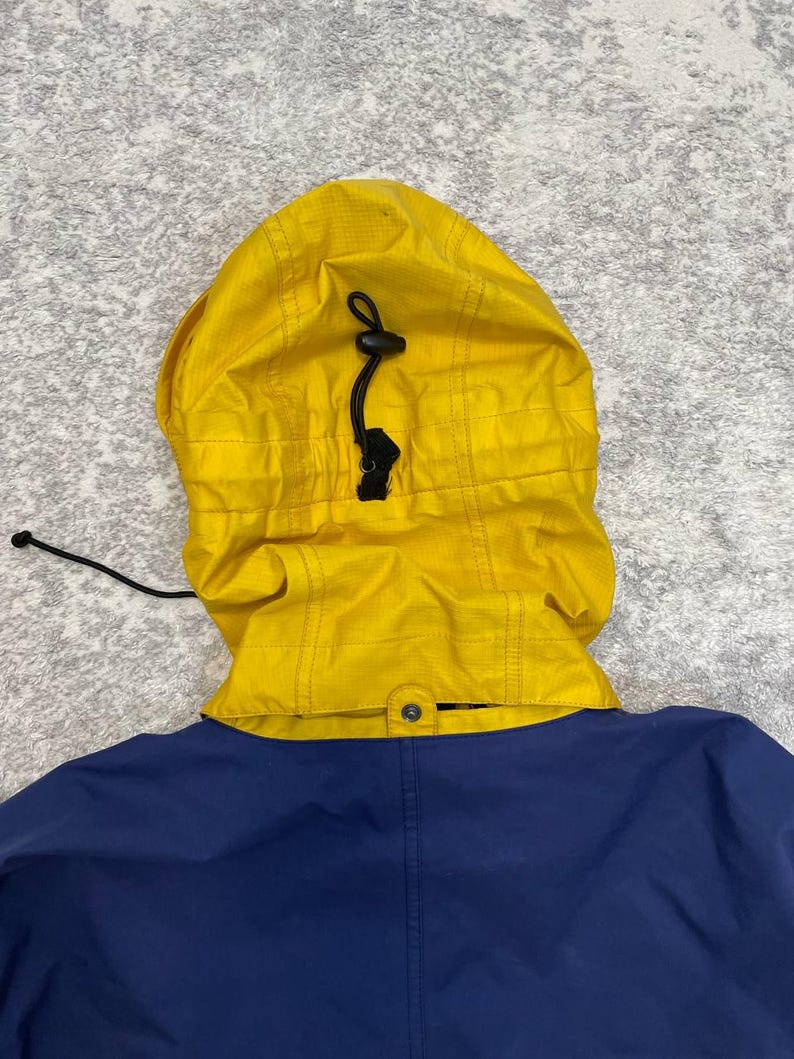 May include: A bright yellow hood with a black drawstring and toggle adjustment attached to a solid navy blue jacket. The hood features a grid-like ripstop texture and is connected to the collar with a single visible snap button at the center.