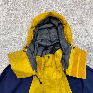 May include: A yellow and navy blue hooded jacket featuring a high collar, a front zipper closure, and a grey interior lining. The word Mammut is printed in red on the lower left side. The hood includes adjustable drawstrings and velcro tabs.