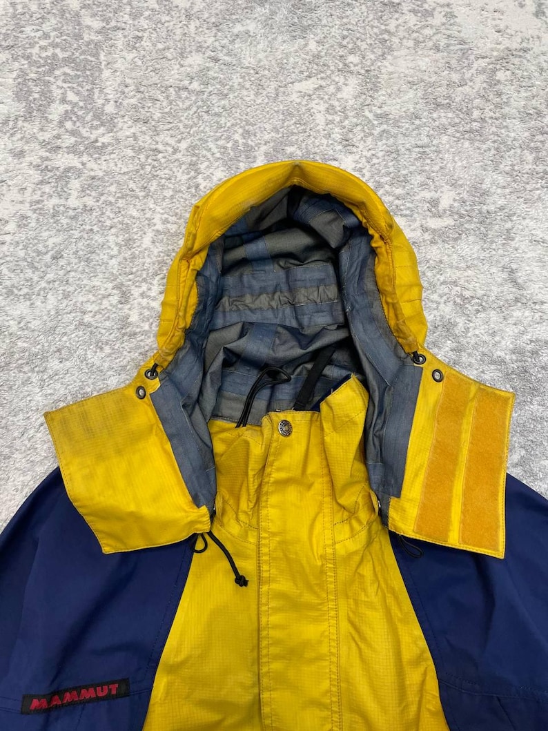 May include: A yellow and navy blue hooded jacket featuring a high collar, a front zipper closure, and a grey interior lining. The word Mammut is printed in red on the lower left side. The hood includes adjustable drawstrings and velcro tabs.