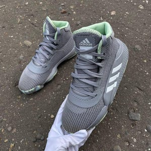 May include: A pair of high-top Adidas basketball sneakers in a grey mesh material with light green interior lining. The shoes feature grey laces, a grey tongue with the Adidas logo, and three white stripes on the side. The soles are grey and light green.