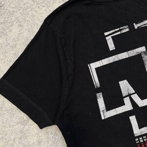 K&ouml;nnte beinhalten: Ein schwarzes kurz&auml;rmeliges T-Shirt mit einem gro&szlig;en, abgenutzten wei&szlig;en Grafikdruck an der Seite. Das Design enth&auml;lt kr&auml;ftige, blockartige Buchstaben und kleineren Text am unteren Rand: PAVOGUR KORINN + IS, STATE PRISON + DK und CZ.