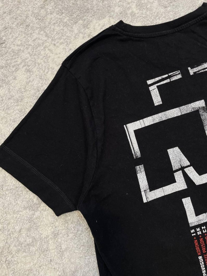K&ouml;nnte beinhalten: Ein schwarzes kurz&auml;rmeliges T-Shirt mit einem gro&szlig;en, abgenutzten wei&szlig;en Grafikdruck an der Seite. Das Design enth&auml;lt kr&auml;ftige, blockartige Buchstaben und kleineren Text am unteren Rand: PAVOGUR KORINN + IS, STATE PRISON + DK und CZ.