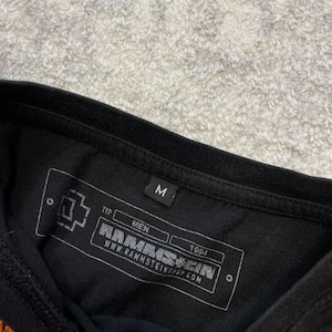 May include: Black t-shirt with a printed label featuring the band name RAMMSTEIN, the year 1994, and the website address. The label also includes the word "MEN" and the letter "M".