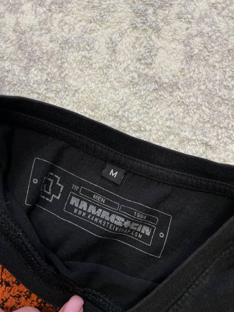 May include: Black t-shirt with a printed label featuring the band name RAMMSTEIN, the year 1994, and the website address. The label also includes the word "MEN" and the letter "M".