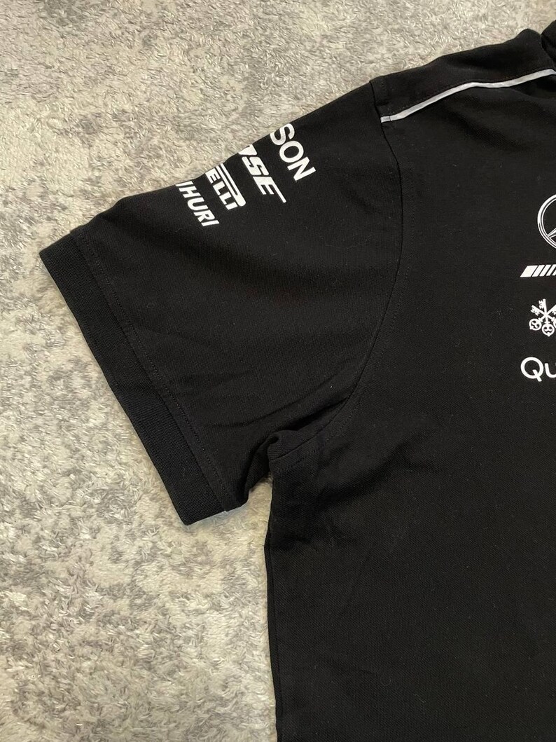 May include: Black short-sleeved t-shirt with white logos on the sleeve and chest. The logos include the words "SON", "SE", "ELLI", and "HURI". The shirt has a crew neck and a white stripe along the shoulder.