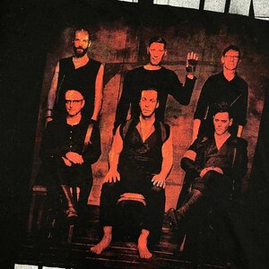 K&ouml;nnte beinhalten: Ein schwarzes T-Shirt mit einem Grafikdruck der Band Rammstein. Das Design zeigt den Bandnamen in wei&szlig;em Text oben, ein Foto der sechs Bandmitglieder in einer dunklen, rot get&ouml;nten Umgebung und den wei&szlig;en Text TOUR 2017 am unteren Rand.