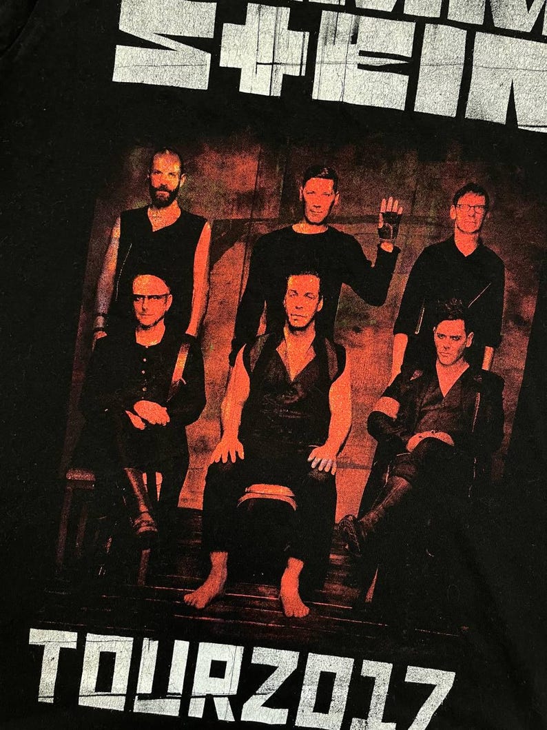 K&ouml;nnte beinhalten: Ein schwarzes T-Shirt mit einem Grafikdruck der Band Rammstein. Das Design zeigt den Bandnamen in wei&szlig;em Text oben, ein Foto der sechs Bandmitglieder in einer dunklen, rot get&ouml;nten Umgebung und den wei&szlig;en Text TOUR 2017 am unteren Rand.