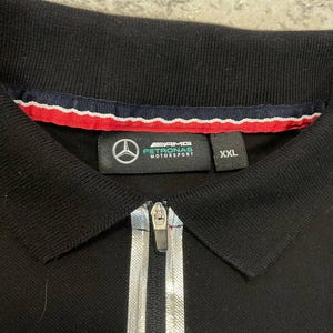 May include: Black polo shirt with a silver zipper and the AMG Petronas Motorsport logo. The shirt has a collar with red, white, and blue trim and a size XXL label.