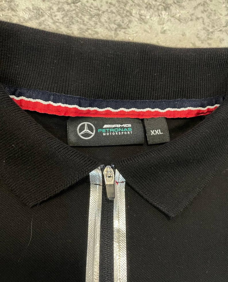 May include: Black polo shirt with a silver zipper and the AMG Petronas Motorsport logo. The shirt has a collar with red, white, and blue trim and a size XXL label.