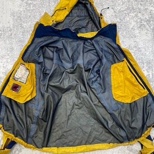 May include: A yellow and navy blue hooded jacket shown from the inside, featuring a grey lining, a hood with adjustable drawstrings, and two large yellow interior pockets. One pocket displays a Mammut logo patch and a Gore-Tex label.