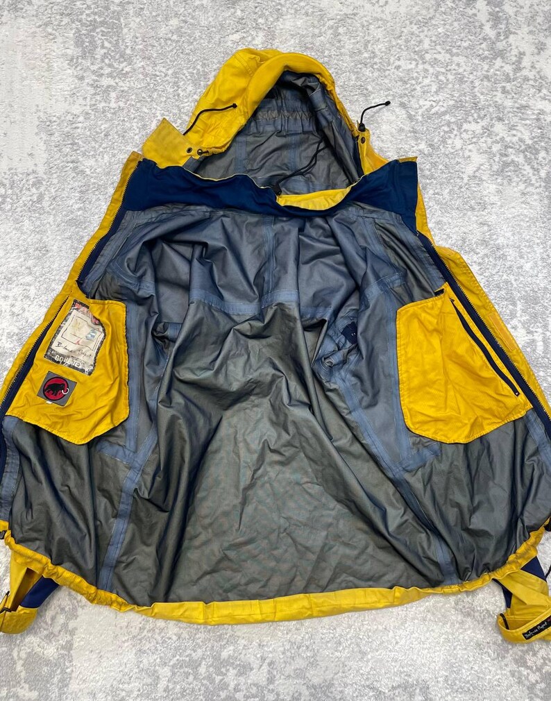 May include: A yellow and navy blue hooded jacket shown from the inside, featuring a grey lining, a hood with adjustable drawstrings, and two large yellow interior pockets. One pocket displays a Mammut logo patch and a Gore-Tex label.