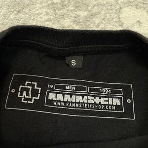 K&ouml;nnte beinhalten: Ein schwarzes T-Shirt mit einem kleinen S-Gr&ouml;&szlig;enetikett am Kragen. Die Innenseite zeigt einen wei&szlig;en, rechteckigen Grafikdruck mit dem Rammstein-Logo, dem Text TYP, MEN, 1994, dem Bandnamen RAMMSTEIN und der Webadresse WWW.RAMMSTEINSHOP.COM.