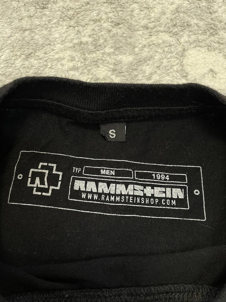 K&ouml;nnte beinhalten: Ein schwarzes T-Shirt mit einem kleinen S-Gr&ouml;&szlig;enetikett am Kragen. Die Innenseite zeigt einen wei&szlig;en, rechteckigen Grafikdruck mit dem Rammstein-Logo, dem Text TYP, MEN, 1994, dem Bandnamen RAMMSTEIN und der Webadresse WWW.RAMMSTEINSHOP.COM.
