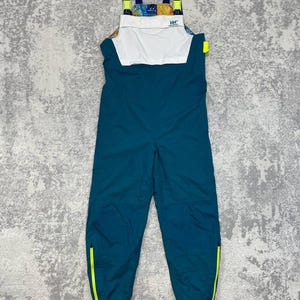 May include: Teal bib overalls featuring a white chest panel with the HH logo, adjustable dark blue shoulder straps with neon yellow accents, and neon yellow zippers at the bottom of each leg. The upper chest area displays a colorful graphic print.