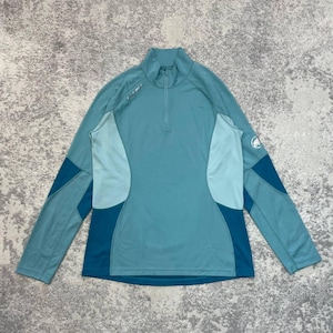 Op de afbeelding: Een blauwgroene trui met lange mouwen, een opstaande kraag en een korte rits. Het kledingstuk is voorzien van kleurvlakken in verschillende tinten blauw en groen, met een wit Mammut-logo op de linkermouw en een klein logo op de borst.
