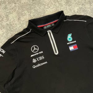 May include: Black polo shirt with a white zipper and collar trim. Features the Mercedes-AMG logo, UBS, and Qualcomm branding, along with a Petronas logo and a flag patch. The shirt has a classic polo collar.