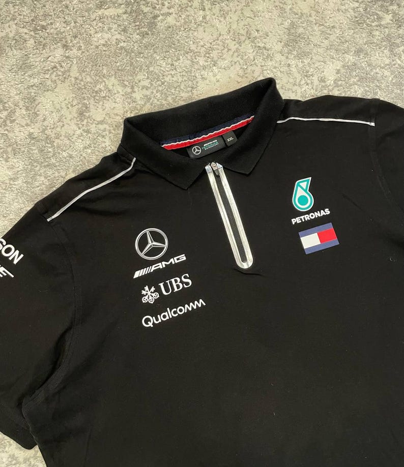 May include: Black polo shirt with a white zipper and collar trim. Features the Mercedes-AMG logo, UBS, and Qualcomm branding, along with a Petronas logo and a flag patch. The shirt has a classic polo collar.
