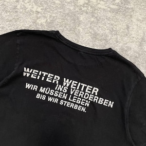 May include: Black t-shirt with white text. The text reads "WEITER WEITER INS VERDERBEN WIR M&Uuml;SSEN LEBEN BIS WIR STERBEN." The shirt is made of a soft material and has a crew neck.