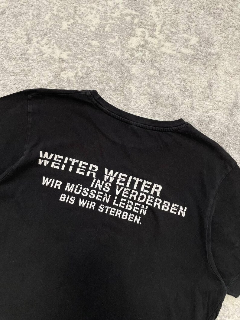 May include: Black t-shirt with white text. The text reads "WEITER WEITER INS VERDERBEN WIR M&Uuml;SSEN LEBEN BIS WIR STERBEN." The shirt is made of a soft material and has a crew neck.