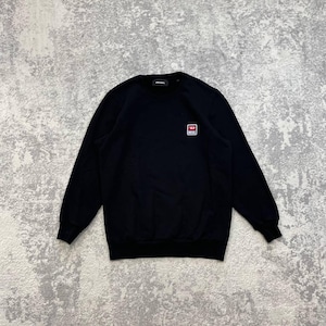 May include: A black long-sleeve crewneck sweatshirt featuring a small rectangular patch on the left chest. The patch displays a red background with a white letter D and the word DIESEL printed in white text below it. The garment has ribbed cuffs.