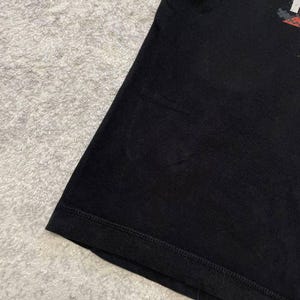 May include: Black t-shirt with a graphic design on the upper right. The shirt is made of a soft material and has a stitched hem. The background is a light gray, textured surface.