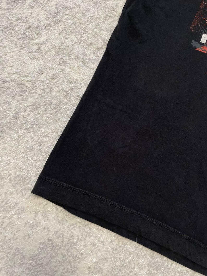 May include: Black t-shirt with a graphic design on the upper right. The shirt is made of a soft material and has a stitched hem. The background is a light gray, textured surface.