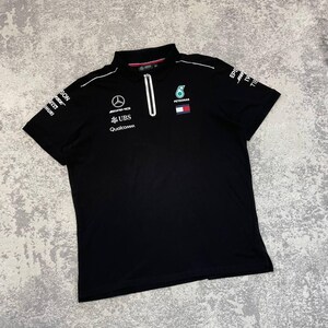 May include: Black polo shirt with a white zipper and collar. The shirt features logos for AMG, UBS, and Qualcomm, along with a Petronas logo. White piping accents the shoulders and sleeves. The shirt is made of a soft, comfortable material.