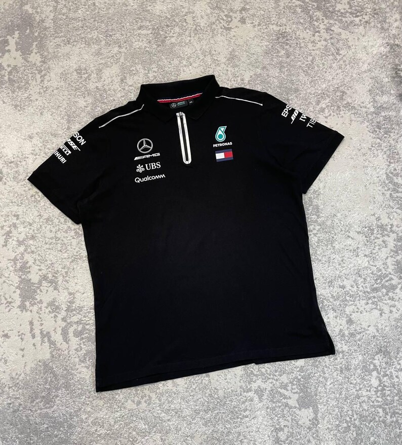 May include: Black polo shirt with a white zipper and collar. The shirt features logos for AMG, UBS, and Qualcomm, along with a Petronas logo. White piping accents the shoulders and sleeves. The shirt is made of a soft, comfortable material.