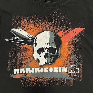 May include: Black t-shirt featuring a graphic design with a skull, an airship, and the band name "RAMMSTEIN" in white and gray. The design includes orange splatter and red accents.