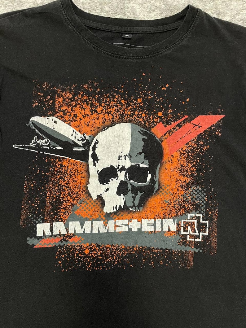 May include: Black t-shirt featuring a graphic design with a skull, an airship, and the band name "RAMMSTEIN" in white and gray. The design includes orange splatter and red accents.