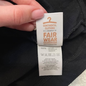 K&ouml;nnte beinhalten: Ein wei&szlig;es Textilpflegeetikett an einem schwarzen Kleidungsst&uuml;ck. Das Etikett zeigt ein orangefarbenes Kleiderb&uuml;gelsymbol, den Text CONTINENTAL CLOTHING IS A MEMBER OF FAIR WEAR FOUNDATION, 100% COTTON sowie Pflegehinweise auf Englisch und Deutsch.