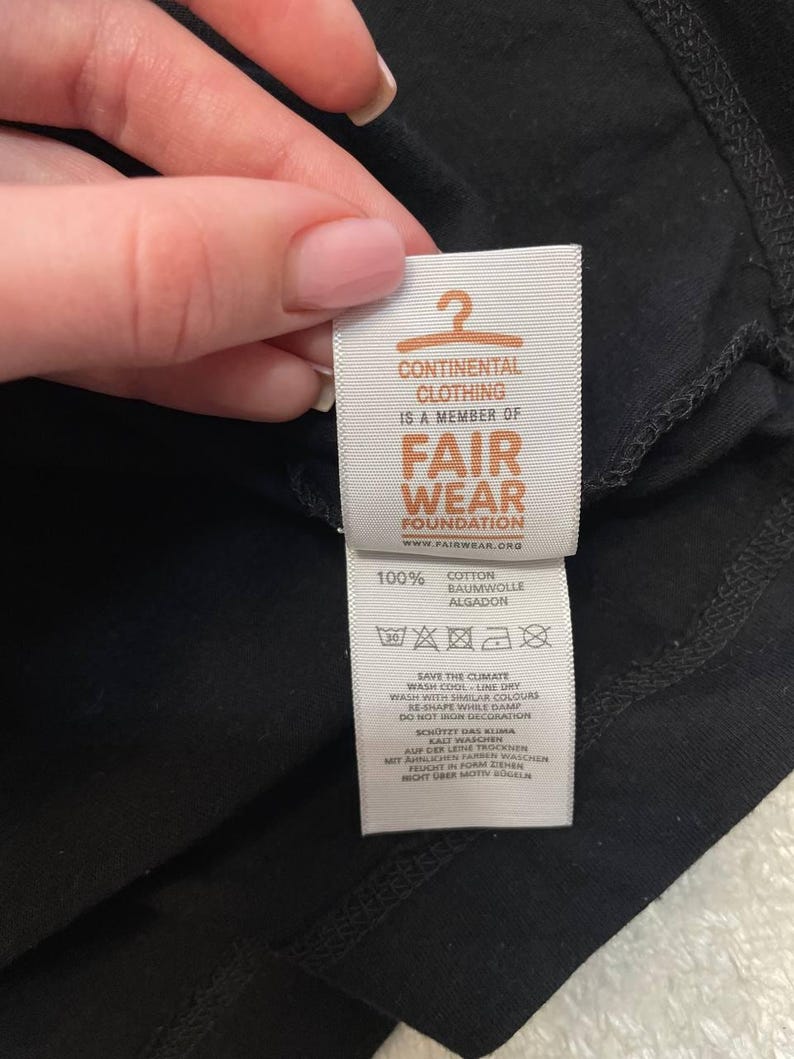 K&ouml;nnte beinhalten: Ein wei&szlig;es Textilpflegeetikett an einem schwarzen Kleidungsst&uuml;ck. Das Etikett zeigt ein orangefarbenes Kleiderb&uuml;gelsymbol, den Text CONTINENTAL CLOTHING IS A MEMBER OF FAIR WEAR FOUNDATION, 100% COTTON sowie Pflegehinweise auf Englisch und Deutsch.