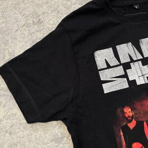 K&ouml;nnte beinhalten: Ein schwarzes Kurzarm-T-Shirt mit einem wei&szlig;en, abgenutzten Grafikdruck im oberen Bereich und einem Foto von zwei Personen darunter. Das T-Shirt hat einen Rundhalsausschnitt und ein kleines schwarzes Etikett mit der Gr&ouml;&szlig;e S am Innenkragen.