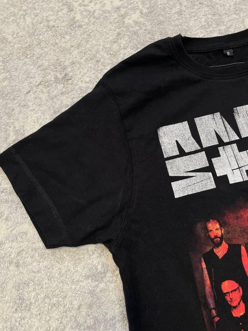 K&ouml;nnte beinhalten: Ein schwarzes Kurzarm-T-Shirt mit einem wei&szlig;en, abgenutzten Grafikdruck im oberen Bereich und einem Foto von zwei Personen darunter. Das T-Shirt hat einen Rundhalsausschnitt und ein kleines schwarzes Etikett mit der Gr&ouml;&szlig;e S am Innenkragen.