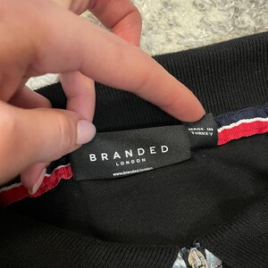May include: Black polo shirt with a zipper and a label that reads "BRANDED LONDON". The label is black with white text and a red, white, and blue stripe detail. The shirt is made in Turkey.