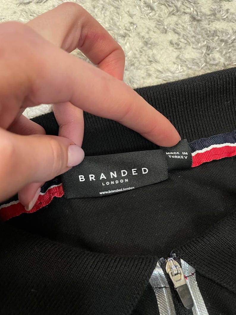 May include: Black polo shirt with a zipper and a label that reads "BRANDED LONDON". The label is black with white text and a red, white, and blue stripe detail. The shirt is made in Turkey.