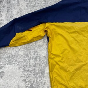 May include: A two-tone jacket featuring a bright yellow lower section and a dark blue upper section. The jacket includes a hood with a black drawstring, a small red brand tag on the sleeve, and a textured fabric finish throughout the garment.