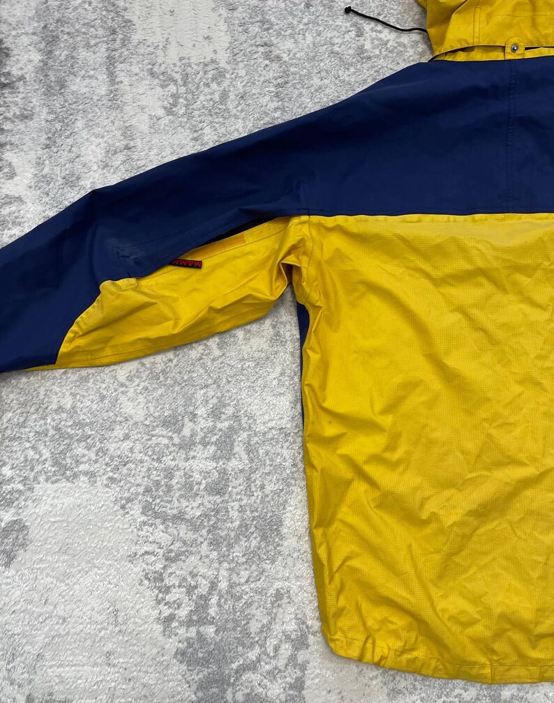 May include: A two-tone jacket featuring a bright yellow lower section and a dark blue upper section. The jacket includes a hood with a black drawstring, a small red brand tag on the sleeve, and a textured fabric finish throughout the garment.